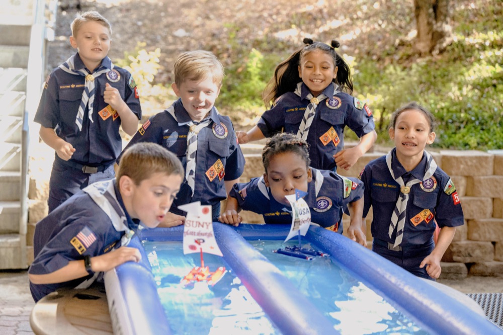Cub Scouts in uniform racing boats down a water-filled trough at a raingutter regatta